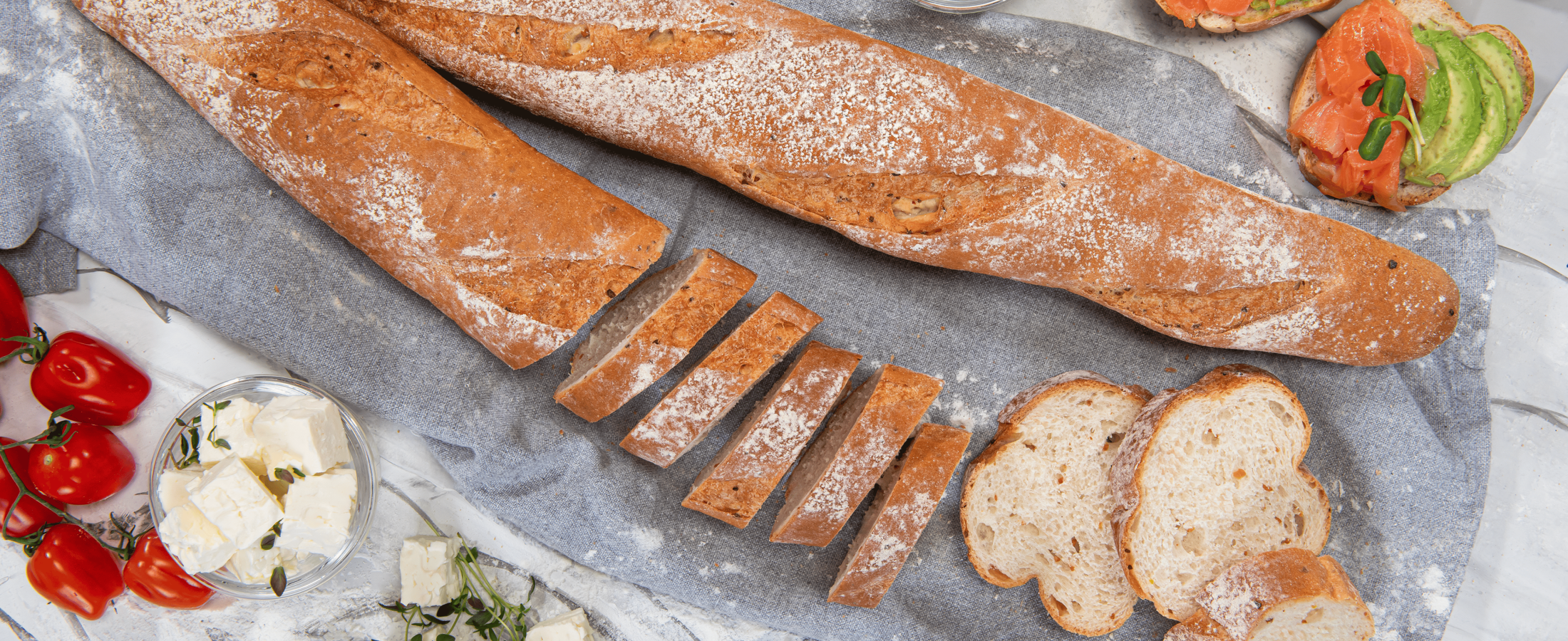 Two rustic baguettes on a cloth, one sliced into pieces, surrounded by tomatoes, cheese cubes, sprouts, and open sandwiches topped with avocado and smoked salmon